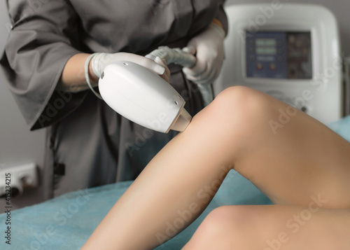 Beautician removing hair on female legs using a laser. Close-up photo of cosmetologist hand with laser maniple. Unfocused laser monitor in the background. Cosmetology and SPA concept. Woman body care.