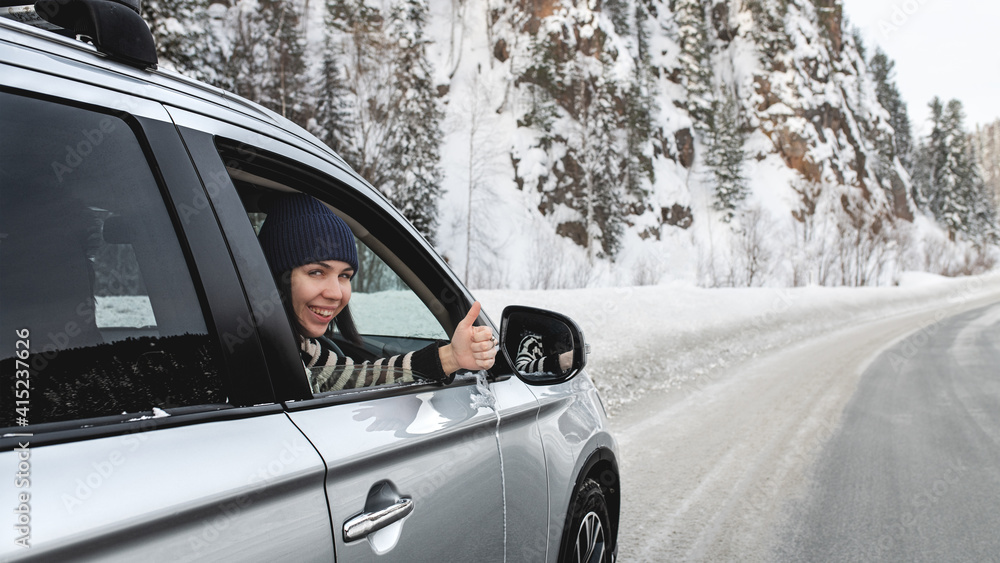 Young pretty brunette sitting in a car and showing thumb up sign through opened side window in winter day on icy road close up.
