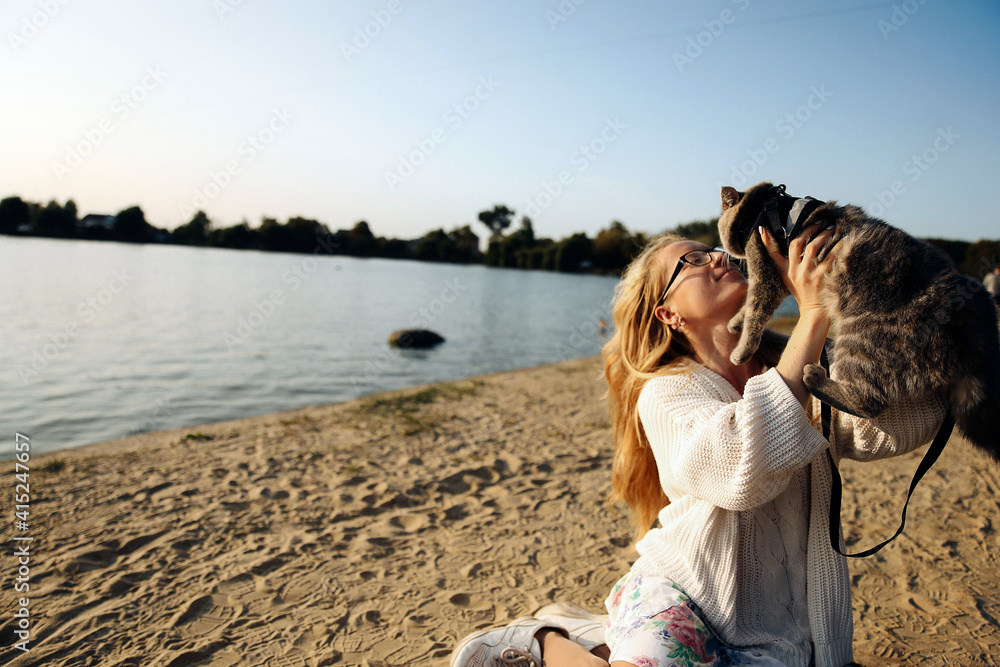 Young blonde woman on the beach in spring in a white knitted sweater with a Scottish straight cat on a sunny day on a picnic