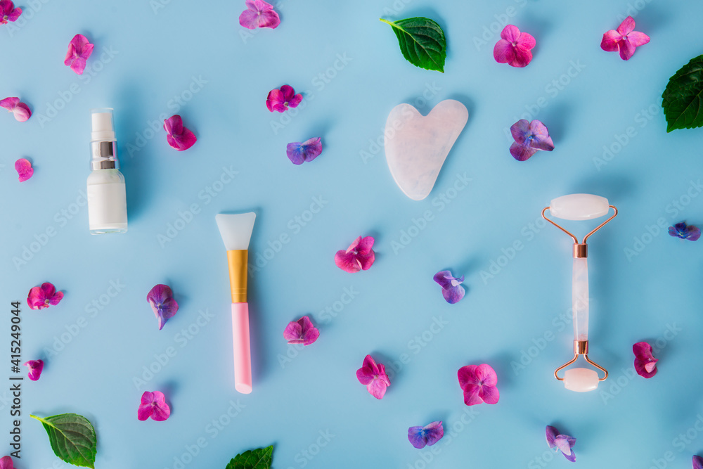 Top view composition with pink jade roller massager, gouache scraper, applying brush and skin serum on blue background with pink hydrangea flower petals. Anti-age, lifting, toning skin treatment. Top view composition with pink jade roller massager, gouache scraper, applying brush and skin serum on blue background with pink hydrangea flower petals. Anti-age, lifting, toning skin treatment.