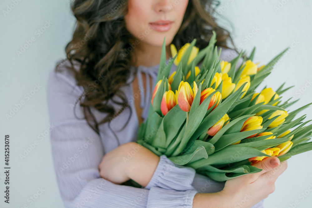 Emotional portrait of young beautiful woman with dark curly hair on light background, holding large bouquet of multicolored tulips in her hands. Spring mood. Mothers Day. Women's Day. Emotional portrait of young beautiful woman with dark curly hair on light background, holding large bouquet of multicolored tulips in her hands. Spring mood. Mothers Day. Women's Day.