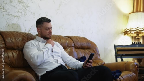 Handsome man on couch with a mobile phone. Young businessman sitting on a sofa in a hotel and unbuttoning his white shirt while viewing in the phone.