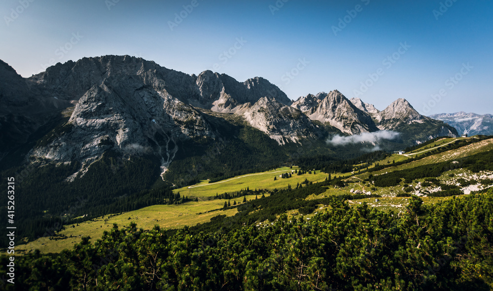 Scenic panoramic view of idyllic rolling hills landscape with blooming meadows and alpine mountain peaks in the background on a beautiful sunny day with blue sky and clouds in summertime