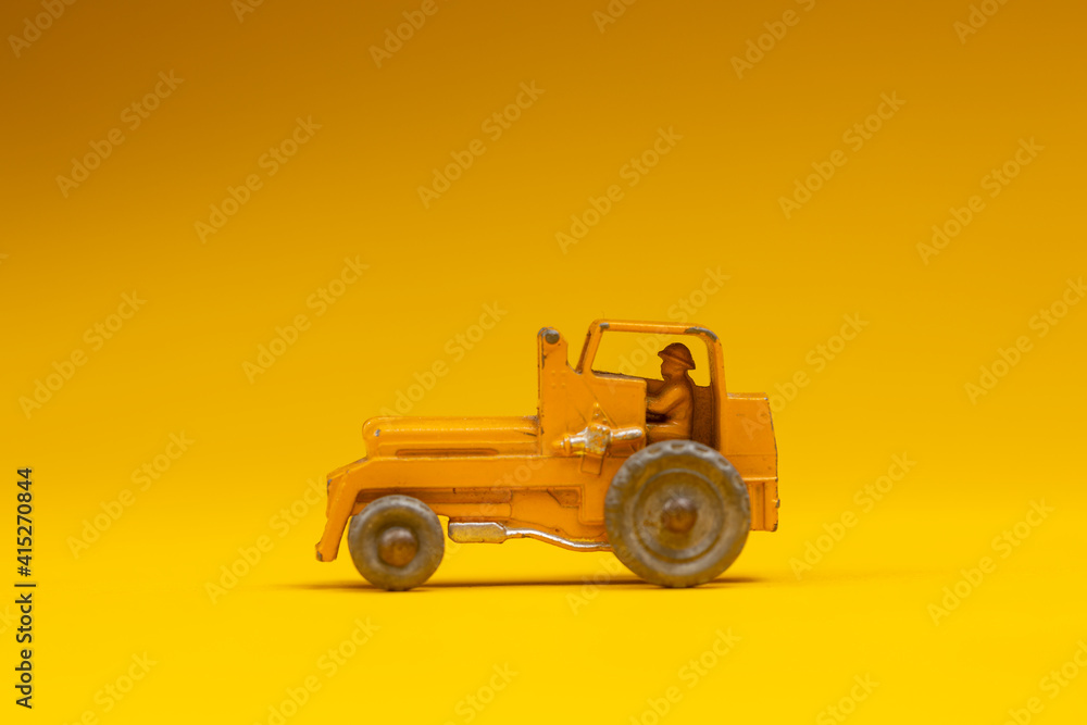 Fototapeta premium Small orange yellow vintage industrial vehicle with figurine inside. Studio toy still life against a seamless yellow background.