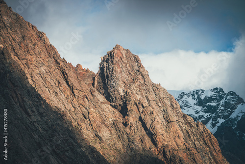 Awesome landscape with sunlit rocky pinnacle on background of high snowy mountains in low clouds. Atmospheric alpine scenery with sharp rock and giant snowy mountain in cloudy sky. Scenic alpine view.