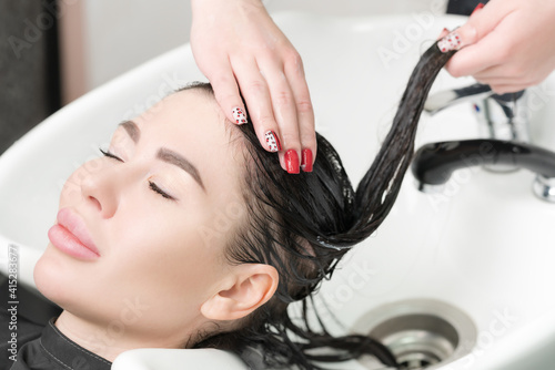 Hairstylist's hands wash long hair of glamour brunette woman with shampoo in professional sink for shampooing in beauty and hair salon.