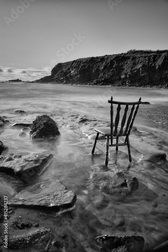 vintage antique chair tucked into the water of an Irish beach surrounded by rocks and cliffs. long exposure with traces of water. mono image photo