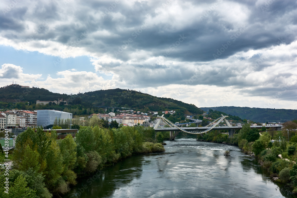 A cityscape with view of the Millenium Bridge on the Minho River seen from the Roman Bridge in the picturesque medieval city of Ourense in Galicia, Spain, on a cloudy spring day.