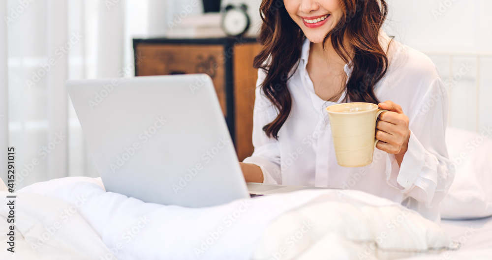 Young smiling happy beautiful asian woman relaxing using laptop computer and drinking coffee in the bedroom at home. Young creative girl working and typing on keyboard.work from home concept Young smiling happy beautiful asian woman relaxing using laptop computer and drinking coffee in the bedroom at home. Young creative girl working and typing on keyboard.work from home concept