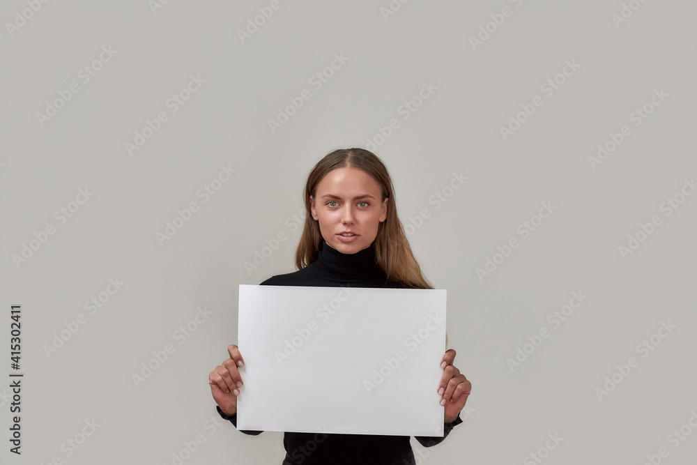 Portrait of attractive young caucasian woman looking at camera, holding white blank banner in front of her while standing isolated over gray background Portrait of attractive young caucasian woman looking at camera, holding white blank banner in front of her while standing isolated over gray background