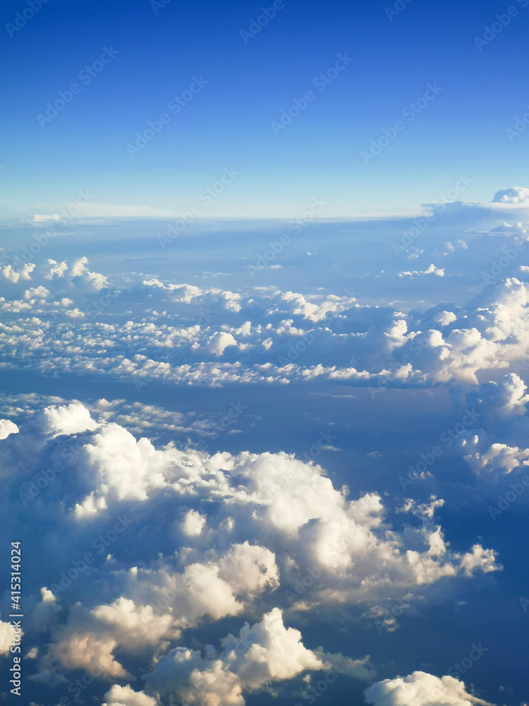 Fototapeta premium A beautiful view from an airplane on white voluminous fluffy clouds against the background of a bright blue sky, city skyscrapers below, top view, vertical frame. Beautiful natural sky background