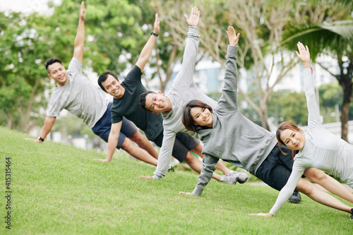 Cheerful young Vietnamese people standing in side plank and looking at camera when working out on campus or in city park © DragonImages
