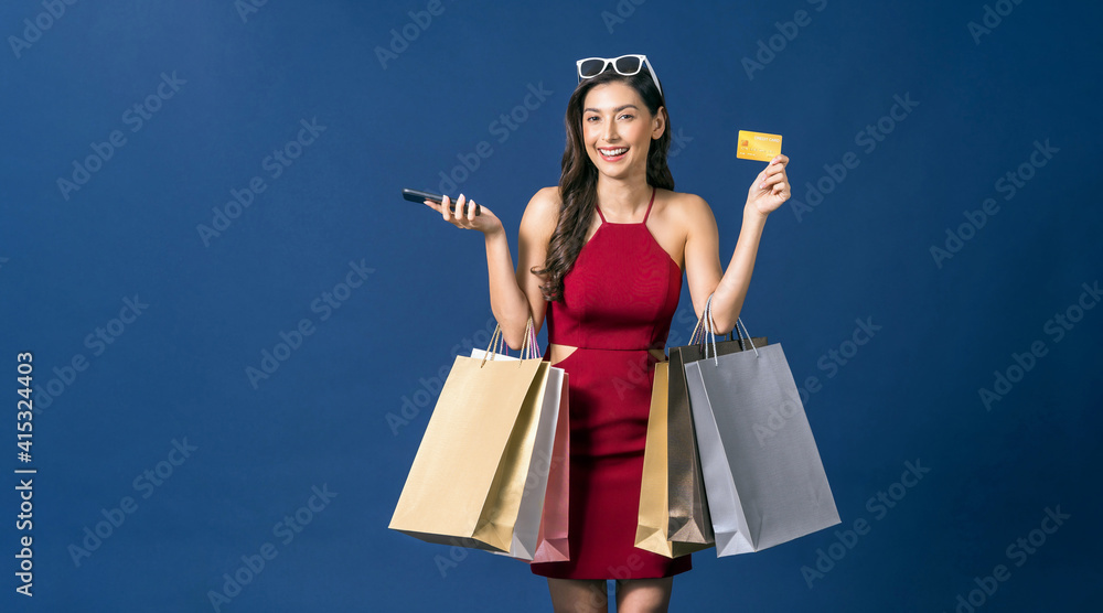 Happy young Asian woman showing credit card and using mobile phone for online shopping on blue color background, cashless payment, black friday and consumerism concept, carrying shopping bags .