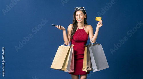 Obraz na płótnie Happy young Asian woman showing credit card and using mobile phone for online shopping on blue color background, cashless payment, black friday and consumerism concept, carrying shopping bags . w salonie