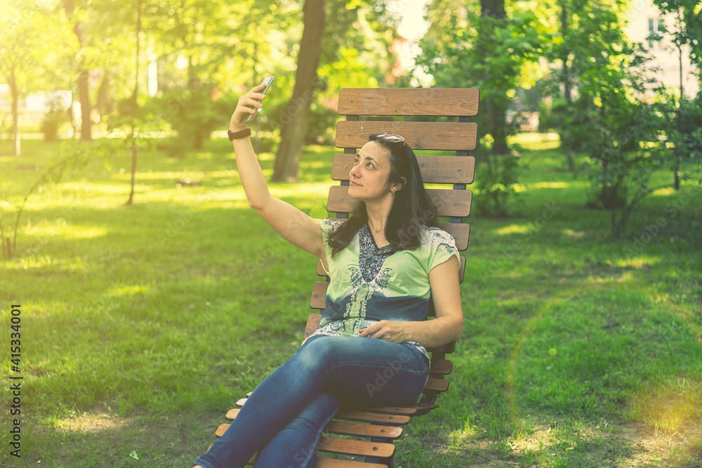 Cheerful girl after shopping with many shopping bags sitting on bench in the park and taking selfie with smart phone. Young woman takes a selfie, sits on a bench in a city park. toned Cheerful girl after shopping with many shopping bags sitting on bench in the park and taking selfie with smart phone. Young woman takes a selfie, sits on a bench in a city park. toned