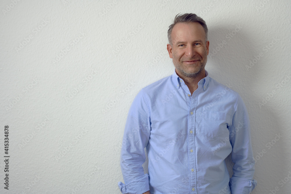 Handsome unshaven mature man in blue shirt, standing against white wall and looking at camera with friendly smile. Half-length front portrait with copy space Handsome unshaven mature man in blue shirt, standing against white wall and looking at camera with friendly smile. Half-length front portrait with copy space