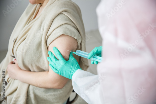 A medical worker in green gloves gives a woman a covid 19 shot in the shoulder. In the frame hands and the syringe