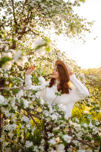Portrait of Beautiful Woman In hat posing near flowering tree. Smiling young woman enjoying smell of flowers on background of spring garden. Fashion, lifestyle. 