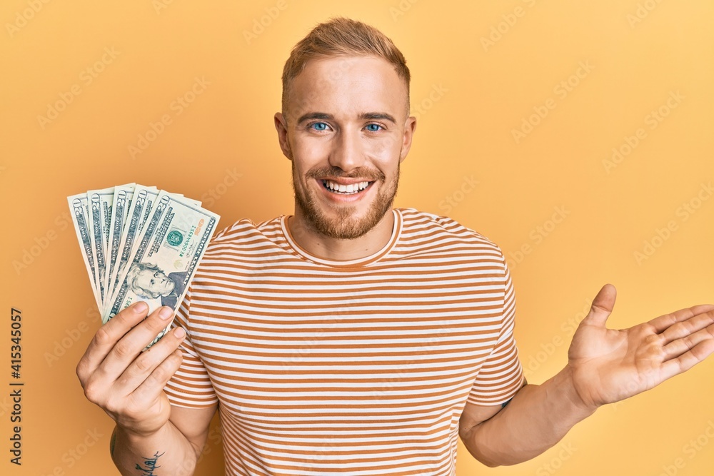 Young caucasian man holding dollars celebrating achievement with happy smile and winner expression with raised hand Young caucasian man holding dollars celebrating achievement with happy smile and winner expression with raised hand