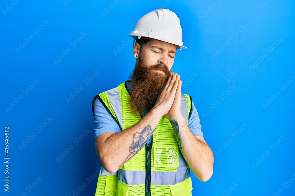 Redhead man with long beard wearing safety helmet and reflective jacket sleeping tired dreaming and posing with hands together while smiling with closed eyes.