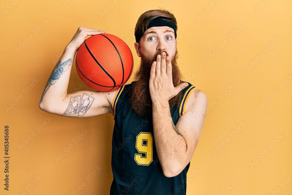 Redhead man with long beard wearing basketball uniform holding ball covering mouth with hand, shocked and afraid for mistake. surprised expression