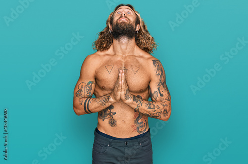 Handsome man with beard and long hair standing shirtless showing tattoos begging and praying with hands together with hope expression on face very emotional and worried. begging.