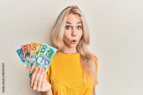 Beautiful caucasian blonde girl holding australian dollars scared and amazed with open mouth for surprise, disbelief face