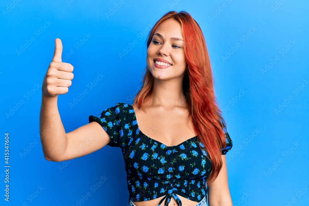 Fototapeta premium Young beautiful redhead woman wearing casual clothes over blue background looking proud, smiling doing thumbs up gesture to the side