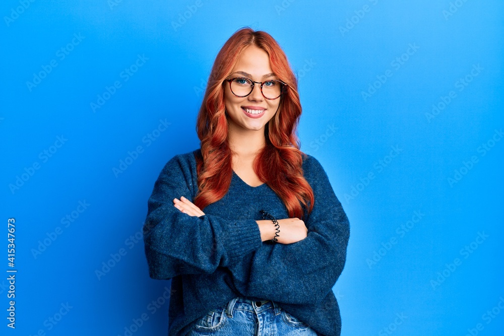 Young beautiful redhead woman wearing casual sweater and glasses over blue background happy face smiling with crossed arms looking at the camera. positive person. Young beautiful redhead woman wearing casual sweater and glasses over blue background happy face smiling with crossed arms looking at the camera. positive person.