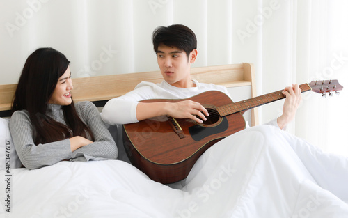 Asian lovers sitting on bed and playing guitar together with fun and happiness. Idea for love and time-sharing of couple