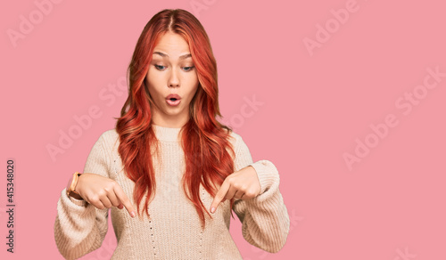Young redhead woman wearing casual winter sweater pointing down with fingers showing advertisement, surprised face and open mouth