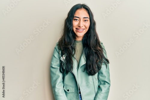 Hispanic teenager girl with dental braces wearing green leather jacket with a happy and cool smile on face. lucky person. photo