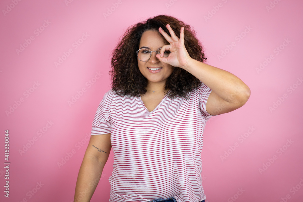 Young african american woman wearing red stripes t-shirt over pink background doing ok gesture shocked with smiling face, eye looking through fingers Young african american woman wearing red stripes t-shirt over pink background doing ok gesture shocked with smiling face, eye looking through fingers