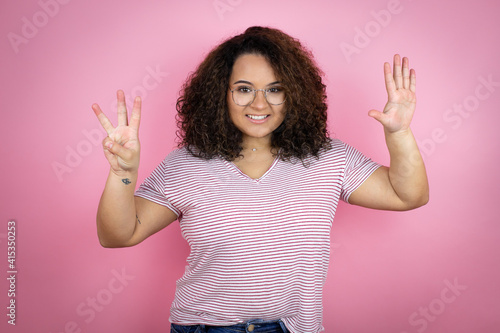 Young african american woman wearing red stripes t-shirt over pink background showing and pointing up with fingers number eight while smiling confident and happy © Irene