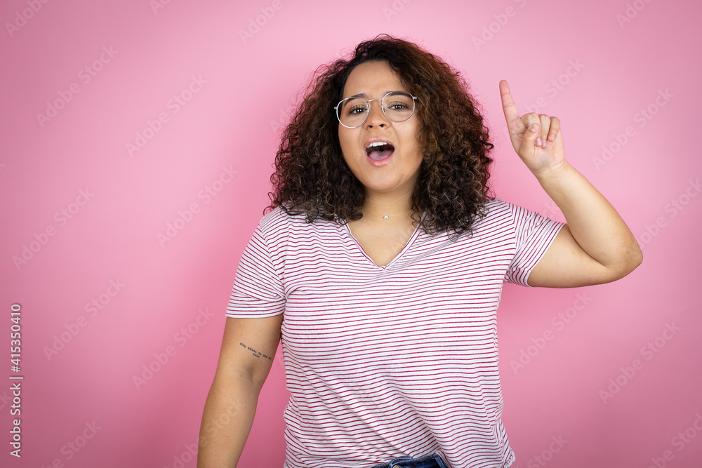 Young african american woman wearing red stripes t-shirt over pink background pointing finger up with successful idea. Exited and happy. Number one. Young african american woman wearing red stripes t-shirt over pink background pointing finger up with successful idea. Exited and happy. Number one.