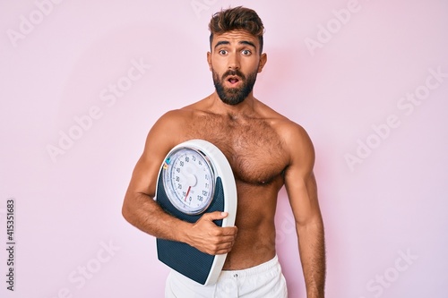 Young hispanic man standing shirtless holding weighing machine scared and amazed with open mouth for surprise  disbelief face