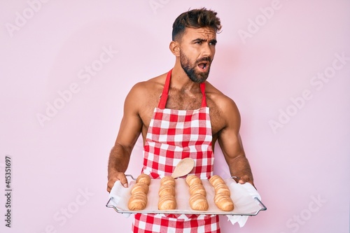 Young hispanic man shirtless wearing baker uniform holding homemade bread angry and mad screaming frustrated and furious, shouting with anger. rage and aggressive concept.