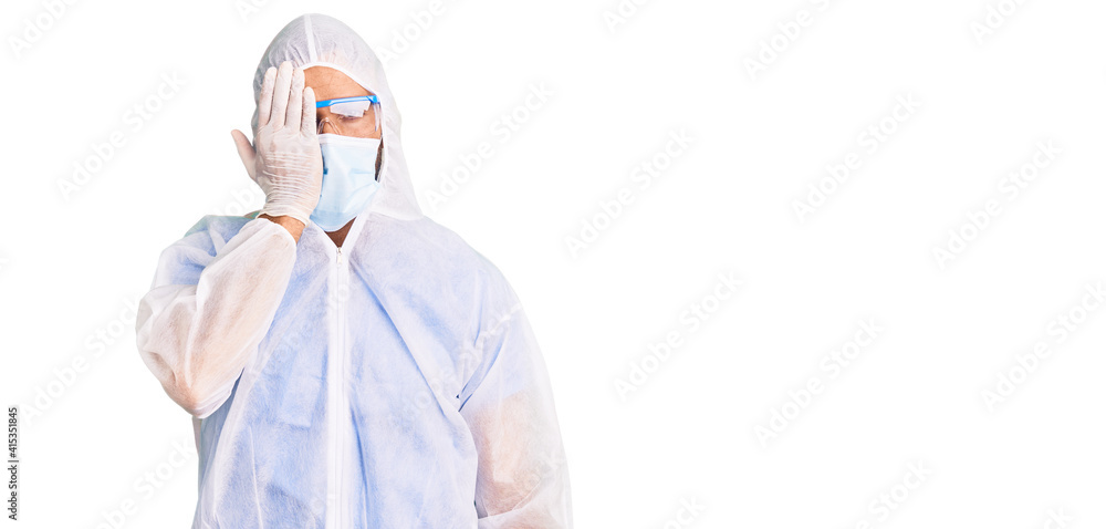 Young hispanic man wearing doctor protection coronavirus uniform and medical mask covering one eye with hand, confident smile on face and surprise emotion. Young hispanic man wearing doctor protection coronavirus uniform and medical mask covering one eye with hand, confident smile on face and surprise emotion.