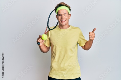 Handsome caucasian man playing tennis holding racket and ball smiling happy and positive, thumb up doing excellent and approval sign © Krakenimages.com
