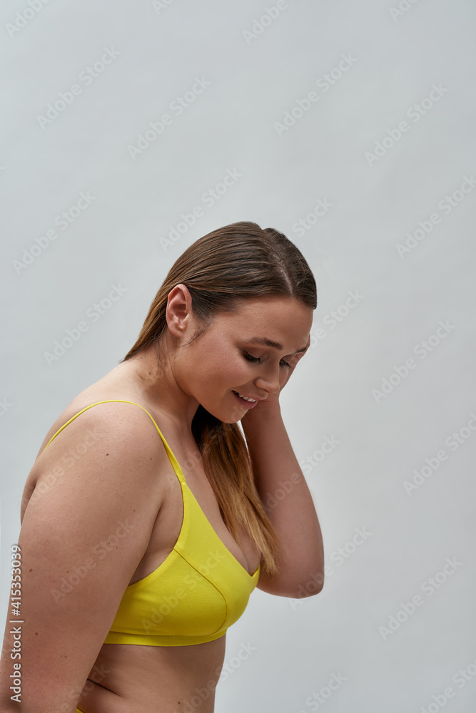 Side view of cute curvy female model wearing yellow underwear smiling while posing isolated over grey background Side view of cute curvy female model wearing yellow underwear smiling while posing isolated over grey background