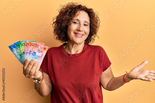 Beautiful middle age mature woman holding swiss franc banknotes celebrating achievement with happy smile and winner expression with raised hand
