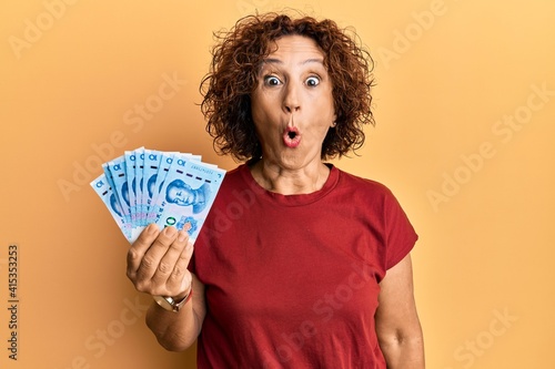 Beautiful middle age mature woman holding 10 yuan chinese banknotes scared and amazed with open mouth for surprise, disbelief face