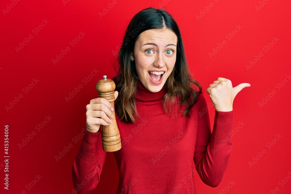 Young brunette woman black pepper grinder pointing thumb up to the side smiling happy with open mouth Young brunette woman black pepper grinder pointing thumb up to the side smiling happy with open mouth