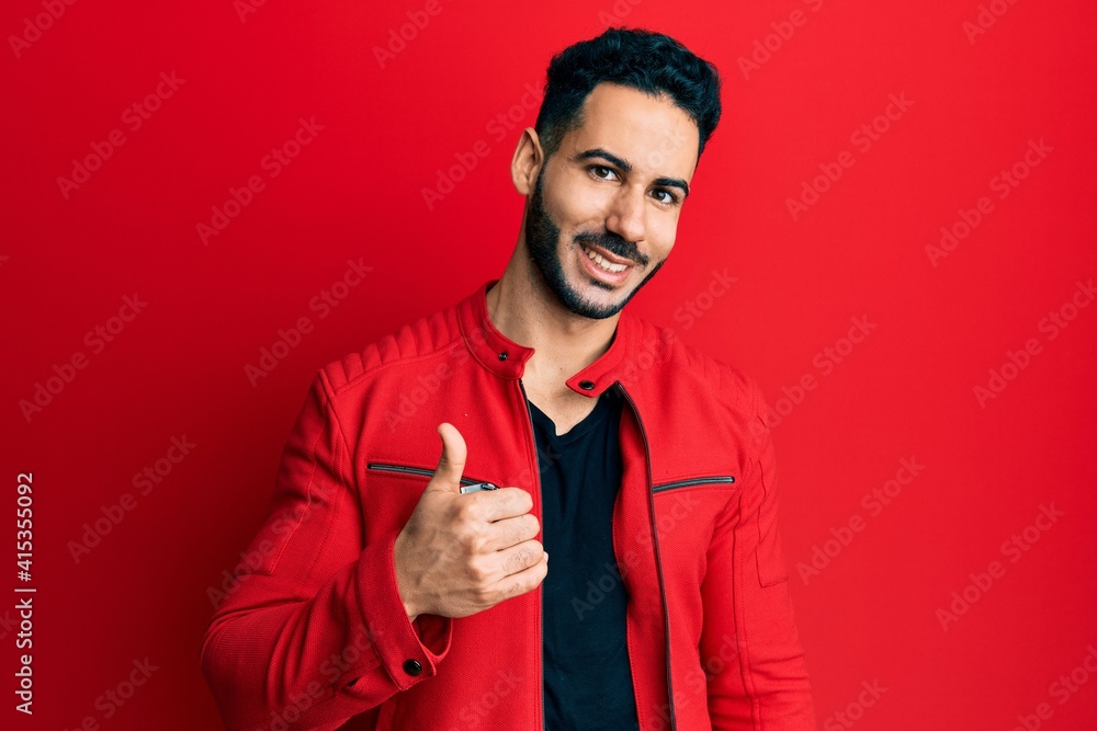 Young hispanic man wearing red leather jacket doing happy thumbs up gesture with hand. approving expression looking at the camera showing success. Young hispanic man wearing red leather jacket doing happy thumbs up gesture with hand. approving expression looking at the camera showing success.