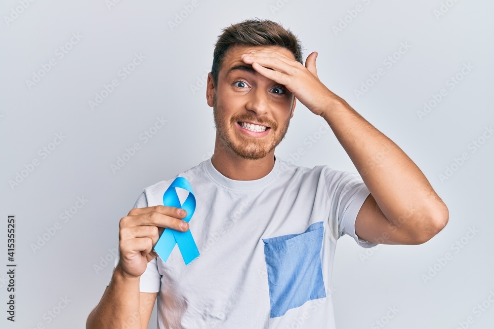 Handsome caucasian man holding blue ribbon stressed and frustrated with hand on head, surprised and angry face Handsome caucasian man holding blue ribbon stressed and frustrated with hand on head, surprised and angry face
