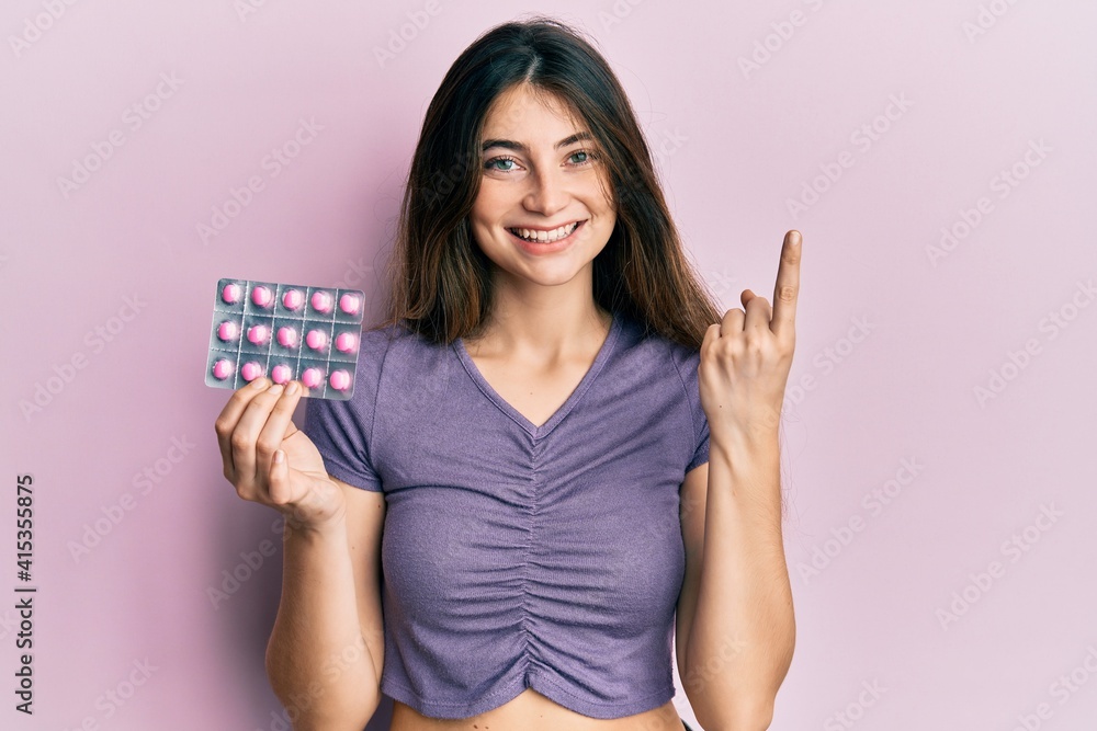Young caucasian woman holding pills smiling with an idea or question pointing finger with happy face, number one