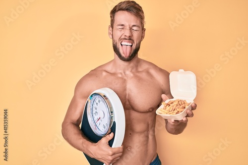 Young caucasian man shirtless holding weighing machine and fried potatoes sticking tongue out happy with funny expression.