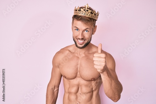 Young caucasian man shirtless wearing king crown smiling happy and positive, thumb up doing excellent and approval sign