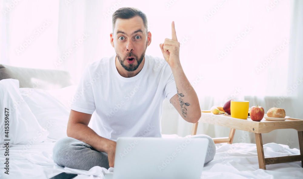 Handsome young modern bearded man works on the laptop while lying on the big white bed at home in the morning Handsome young modern bearded man works on the laptop while lying on the big white bed at home in the morning