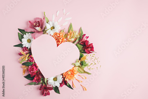 Heart surrounded with fresh colorful flowers on pastel pink background. Creative love layout with copy space. Valentines day, wedding or romantic visual trend. Spring bloom. Flat lay.